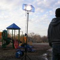 Two tall and bright hot lamps are standing on a playground. The play place is in the background. One man stands in the foreground, facing away from camera